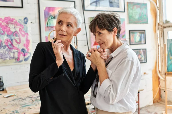 Fashionable lesbian couple holding hands and looking around in workshop with creative paintings — Stock Photo