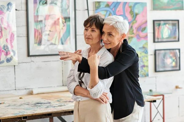 Stylish mature woman embracing lesbian partner and pointing with hand in modern art workshop — Stock Photo