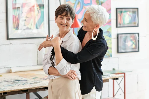 Cheerful and trendy mature woman embracing lesbian partner in modern art workshop, happiness — Stock Photo