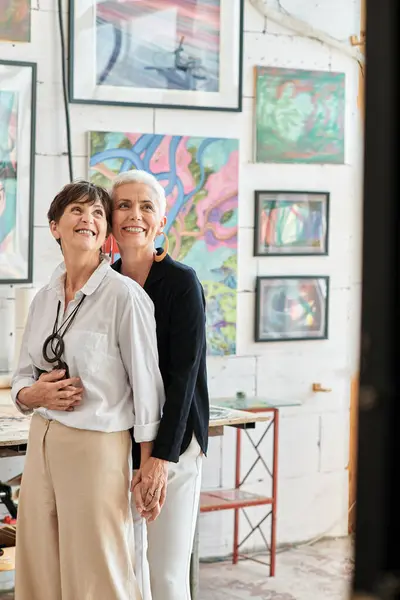 Alegre y elegante lesbianas artistas abrazando y mirando hacia otro lado en el taller de arte moderno, juntos - foto de stock