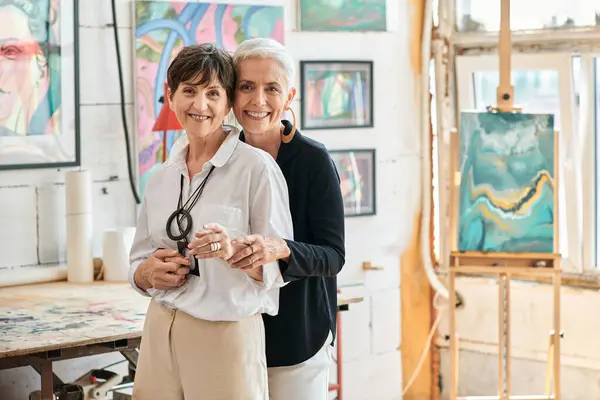 Des artistes lesbiennes souriants embrassant et regardant la caméra dans un atelier d'art moderne, ensemble — Photo de stock