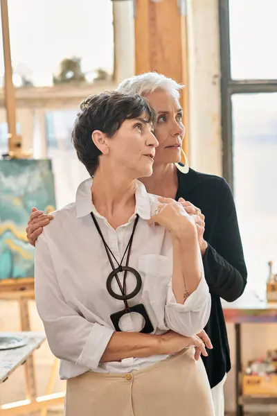 Artistes lesbiennes réfléchies et élégantes regardant loin dans le studio d'art moderne avec des peintures créatives — Photo de stock