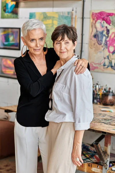 Elegant and happy lesbian couple of modern artists standing and looking away in craft workshop — Stock Photo