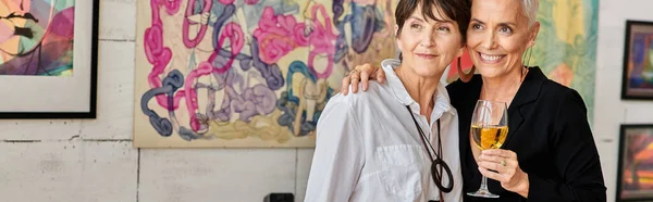 Smiling mature woman with wine glass embracing lesbian partner in modern art studio, banner — Stock Photo