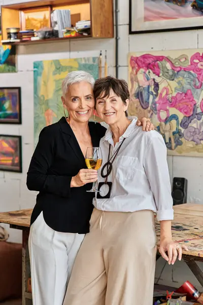 Lesbian couple of artists with wine glass embracing and looking at camera in contemporary art studio — Stock Photo