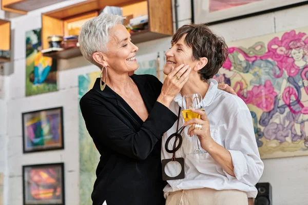 Trendige reife Frau berührt Gesicht lesbischer Partnerin mit Weinglas im Studio für zeitgenössische Kunst — Stockfoto