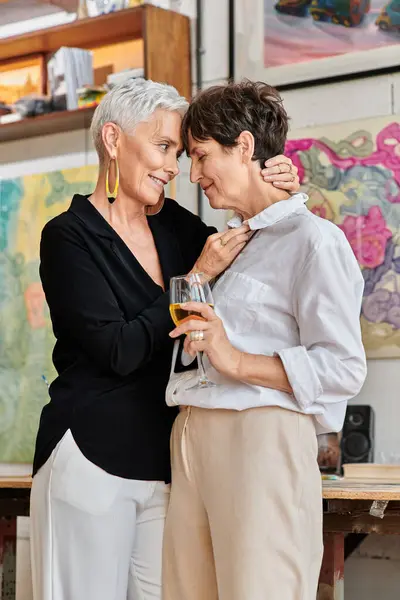 Stylish middle aged woman embracing lesbian partner with wine glass in contemporary art studio — Stock Photo