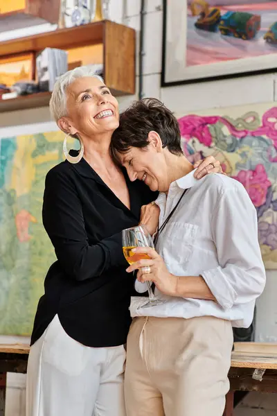 Aufgeregte Frau mittleren Alters umarmt lesbische Partnerin mit Weinglas in modernem Kunstworkshop — Stockfoto