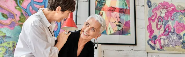 Elegantes y alegres lesbianas maduras artistas sonriendo entre sí en el taller de arte contemporáneo, pancarta - foto de stock