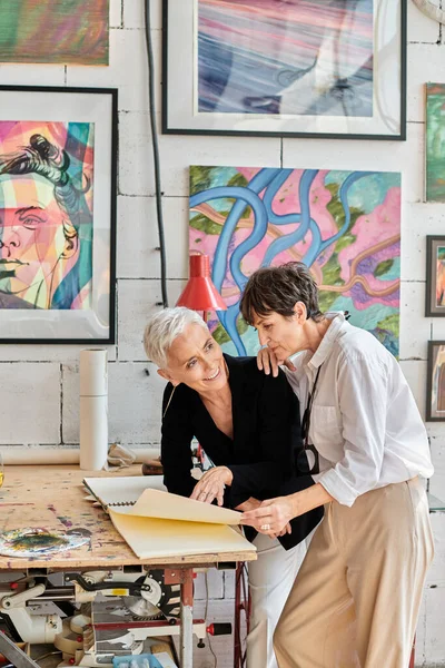 Artistas lésbicas talentosos e felizes olhando através do álbum no estúdio de arte com pinturas coloridas — Fotografia de Stock