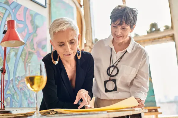 Artistes lesbiennes élégants et joyeux souriant près de l'album dans un studio d'art avec des peintures colorées — Photo de stock