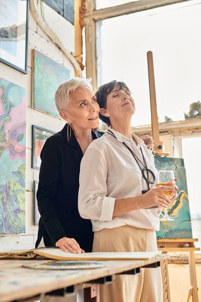 Зрелая женщина с закрытыми глазами и бокалом вина возле стильного лесбийского партнера в студии современного искусства — стоковое фото