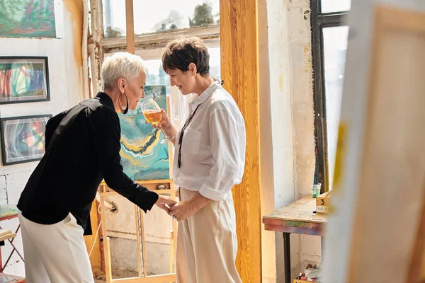 Artiste souriante tenant verre à vin près partenaire féminine dans un studio d'art moderne, ensemble — Photo de stock