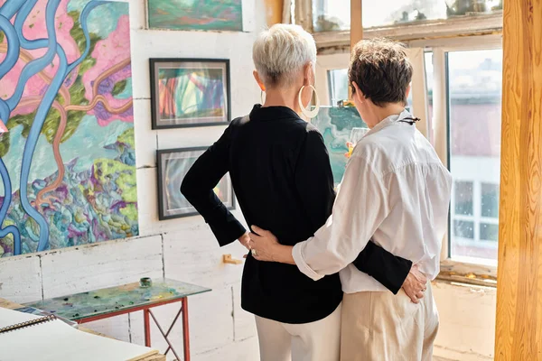 Visão traseira de casal lésbico elegante de artistas abraçando e de pé na oficina de arte moderna — Fotografia de Stock