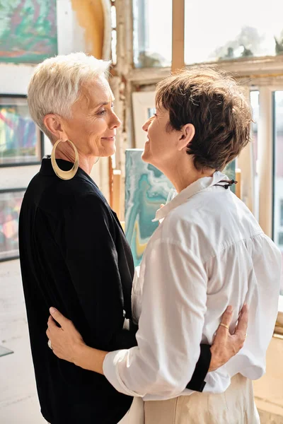 Веселые и элегантные зрелые лесбийские художники, смотрящие друг на друга в студии современного искусства — стоковое фото