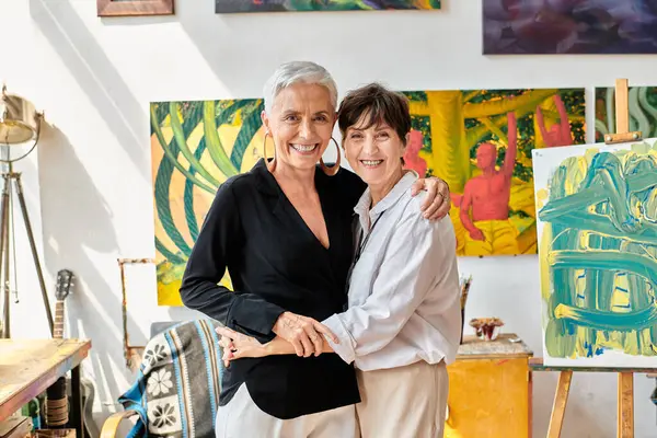Cheerful mature lesbian artists embracing and smiling at camera in contemporary art workshop — Stock Photo