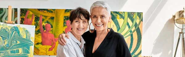 Joyful mature lesbian artists embracing and looking at camera in contemporary art workshop, banner — Stock Photo