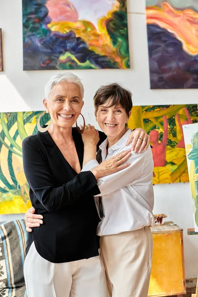 Cheerful mature lesbian artists embracing and smiling at camera near colorful paintings in workshop — Stock Photo