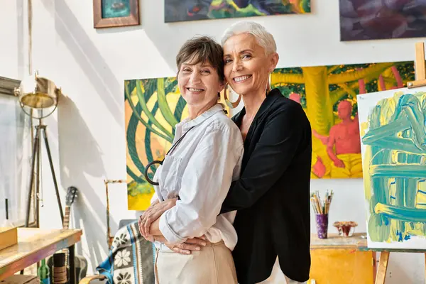 Peintres lesbiennes branchés et heureux embrassant et regardant la caméra près de peintures créatives en studio — Photo de stock