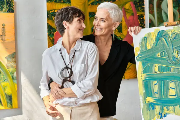 Talentueux artistes lesbiennes matures embrassant et souriant à l'autre près de peintures dans l'atelier — Photo de stock