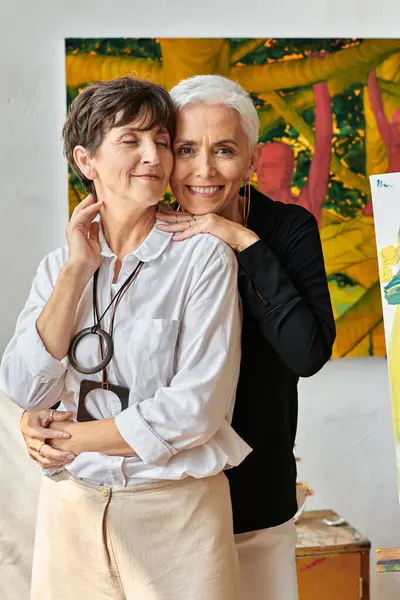 Talented middle aged lesbian artists embracing near creative paintings in modern craft workshop — Stock Photo