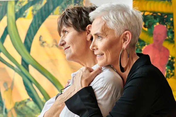 Vue latérale des artistes matures lesbiennes à la mode et souriantes regardant loin dans l'atelier d'art contemporain — Photo de stock