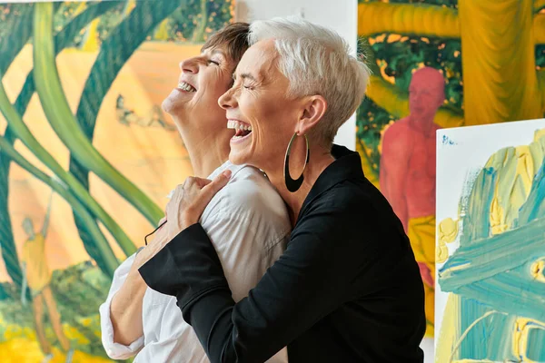 Side view of excited and stylish lesbian artists laughing with closed eyes in modern craft workshop — Stock Photo