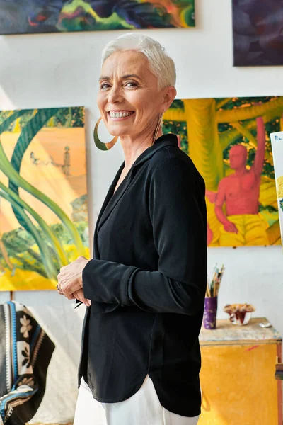 Стильная и веселая женщина средних лет, смотрящая на камеру рядом с творческими картинами в студии — стоковое фото