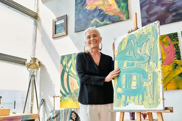 Elegant and excited female artist smiling at camera near easel and colorful paintings in studio — Stock Photo