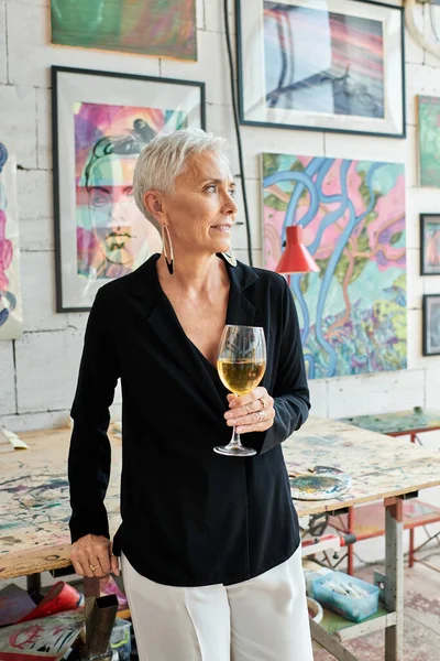 Soñadora madura artista femenina con copa de vino mirando hacia otro lado en el estudio de arte con pinturas creativas - foto de stock