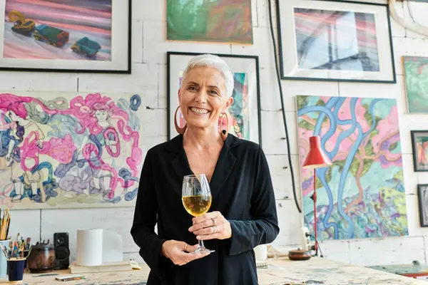 Alegre madura artista femenina con copa de vino sonriendo a la cámara en el estudio de arte con pinturas creativas - foto de stock