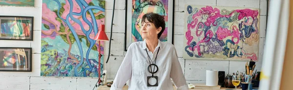 Nachdenkliche reife Künstlerin steht und schaut weg in Kunstatelier mit bunten Gemälden, Banner — Stockfoto
