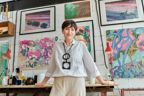 Artista mulher alegre e elegante olhando para a câmera no estúdio de arte com pinturas expressivas coloridas — Fotografia de Stock