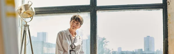 Smiling mature female artist looking at camera near window modern spacious workshop, banner — Stock Photo