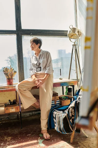 Alegre artista femenina madura sentada cerca de la ventana y mirando hacia otro lado en moderno y espacioso taller - foto de stock
