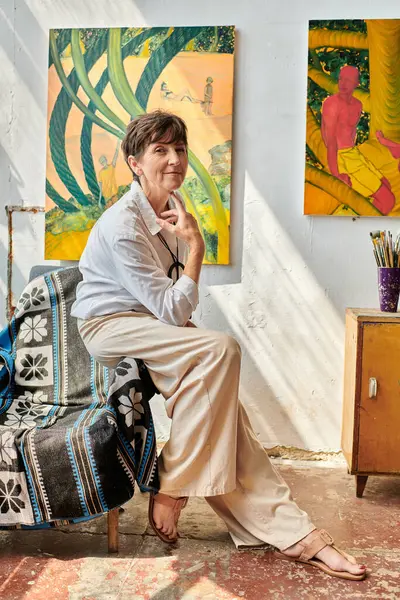 Femme d'âge moyen à la mode artiste assis sur un fauteuil et souriant à la caméra dans un studio spacieux moderne — Photo de stock