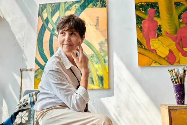 Мечтательная и улыбающаяся художница средних лет, сидящая и смотрящая в мастерскую современного искусства — стоковое фото