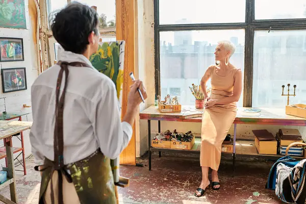 Mulher de meia idade em vestido elegante posando perto artista feminino em oficina leve e espaçosa — Fotografia de Stock