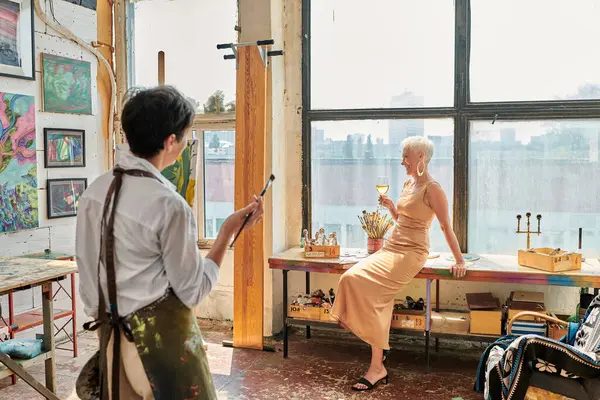 Стильная зрелая женщина позирует с бокалом вина рядом с художницей в светлой и просторной мастерской — стоковое фото