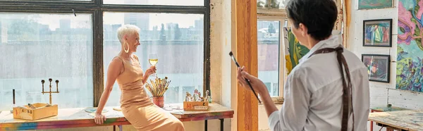 Modelo maduro elegante posando com copo de vinho perto artista mulher na oficina moderna, bandeira horizontal — Fotografia de Stock