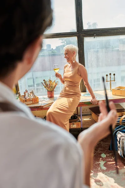 Joyful mature model posing with wine glass near blurred woman artist in modern art workshop — Stock Photo