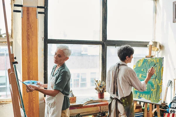 Креативні жінки середнього віку в фартухах живопис на мольбертах в сучасній майстерні ремесел, художнє хобі — стокове фото