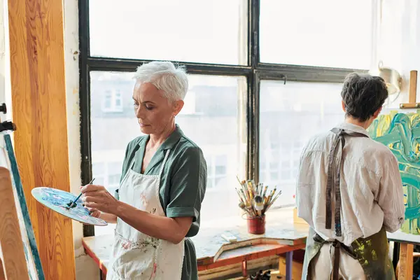 Concentrated mature woman with palette near female friend painting in art studio, creative hobby — Stock Photo
