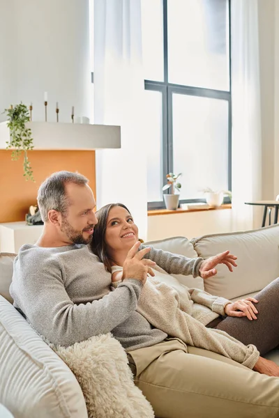Alegre pareja sin niños sentados en un cómodo sofá y hablando en la moderna sala de estar en casa - foto de stock