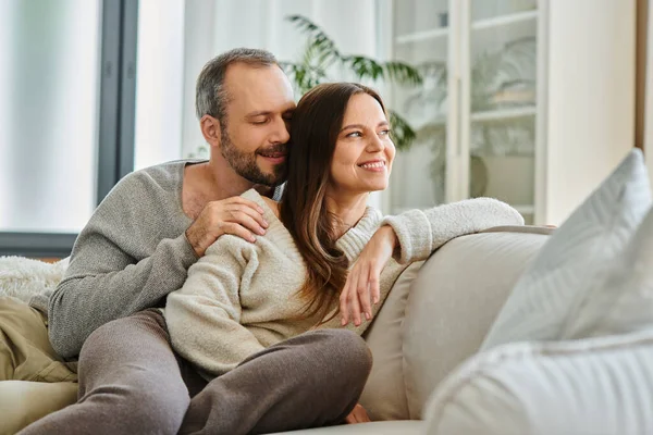 Sonriente hombre con los ojos cerrados abrazando esposa en cómodo sofá en la sala de estar, pareja libre de niños - foto de stock