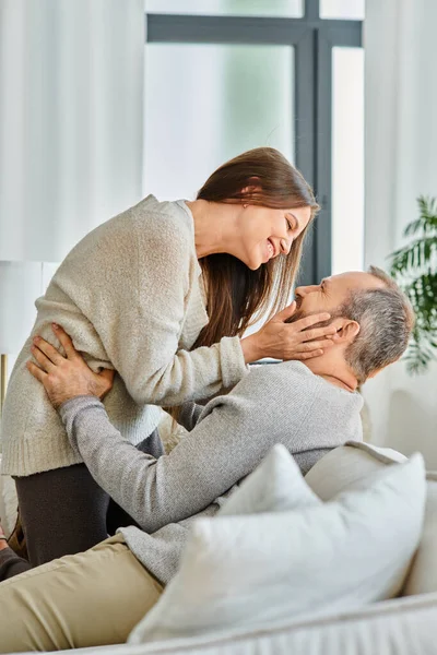Mujer sonriente abrazando la cara de marido feliz en la sala de estar en casa, ocio de pareja libre de niños - foto de stock