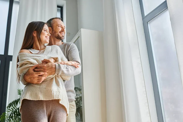 Joyful child-free couple embracing and looking away near window in living room, tranquil lifestyle — Stock Photo