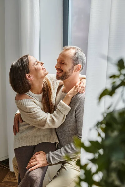 Laughing child-free couple embracing and smiling at each other near window at home, tenderness — Stock Photo
