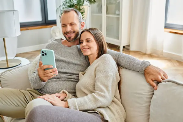 Sonriente hombre navegar por Internet en el teléfono inteligente cerca de la esposa en el sofá en la sala de estar, pareja libre de niños - foto de stock