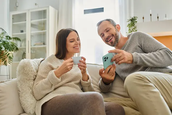 Збуджений чоловік показує смартфон дружині з чашкою кави на дивані у вітальні, бездітна пара — стокове фото